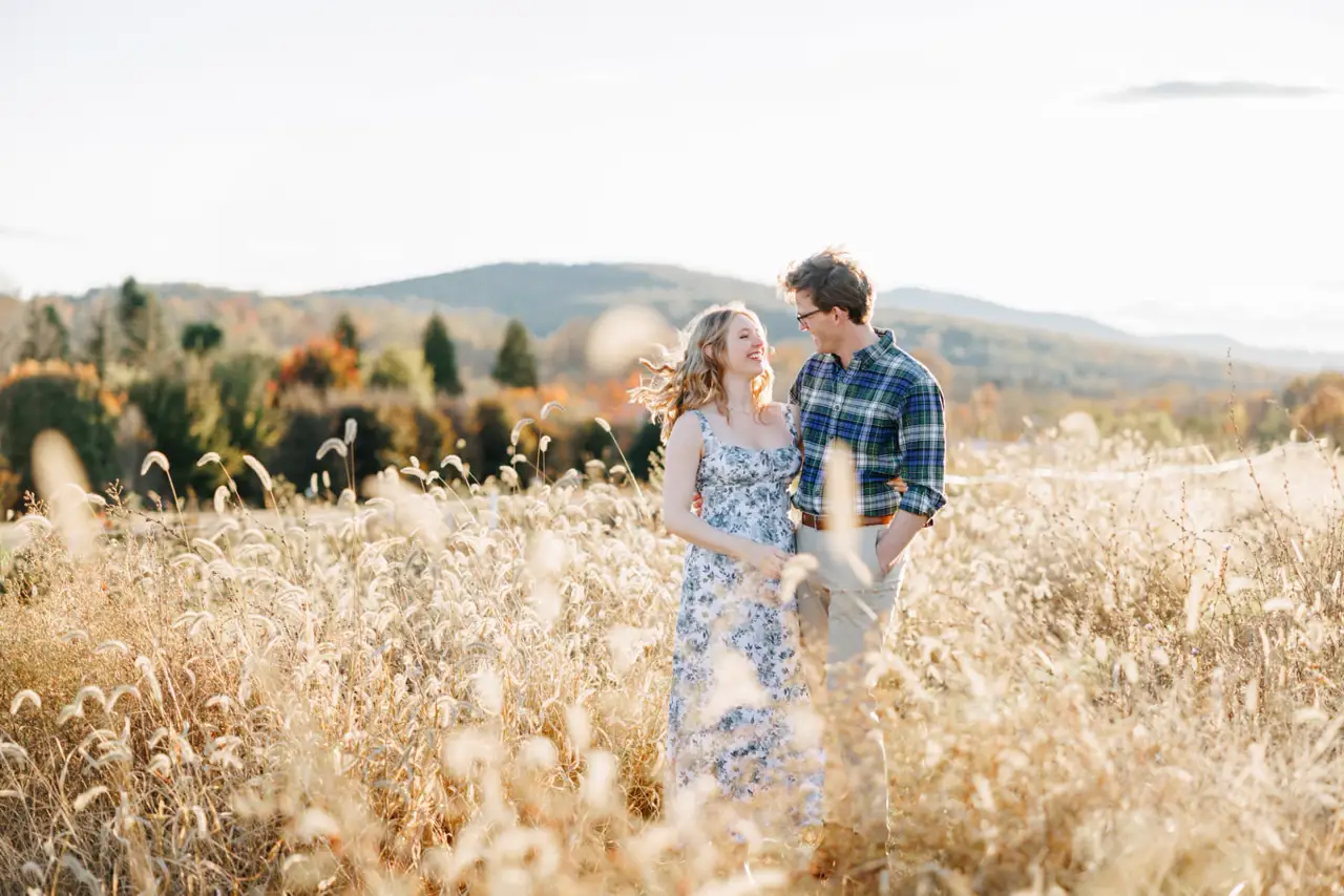 An engaged couple laughing together in a golden field with mountain views at golden hour